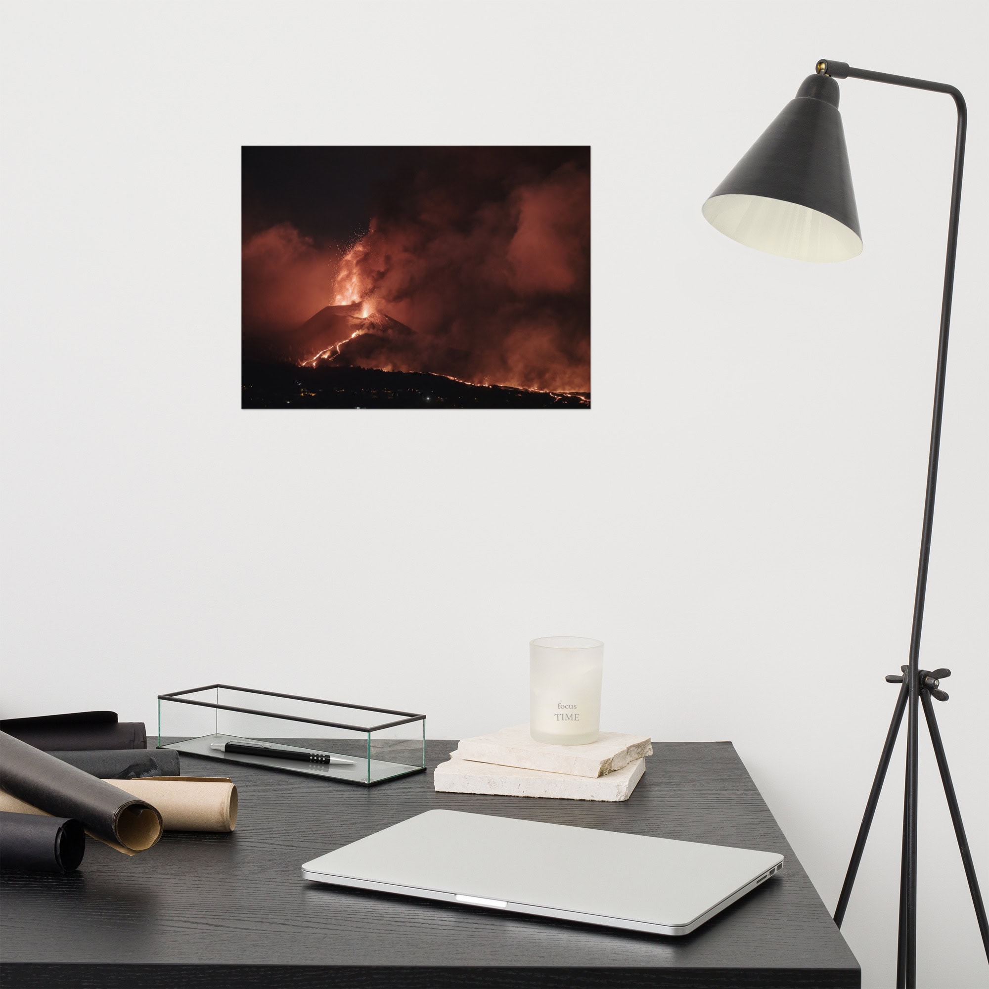 Un espacio de trabajo minimalista muestra un escritorio negro adornado con una computadora portátil cerrada, dos papeles negros enrollados, una vela y un marco de fotos. Una lámpara en blanco y negro está ubicada al lado del escritorio. En la pared cuelga un impresionante Póster volcán 2021 La Palma noche, que captura la ardiente erupción volcánica por la noche y agrega un toque de arte decorativo.