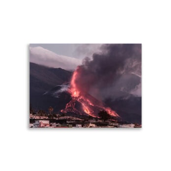 Un volcán entra en erupción y cae lava de un rojo intenso por sus laderas, lanzando densas columnas de humo oscuro al cielo. En primer plano aparecen edificios residenciales bajo un cielo nublado, una imagen perfectamente captada por el Póster volcán 2021 La Palma edificios de Volcanica Productions.