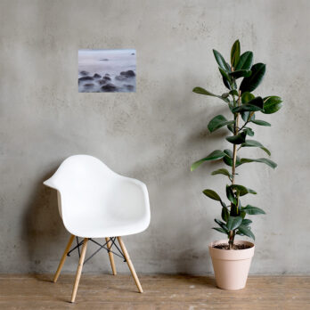 Una elegante silla blanca se encuentra junto a una maceta alta con hojas verdes anchas, contra una pared gris minimalista. Sobre la silla, una pequeña obra de arte titulada "Póster de Rocas de la playa de El Lloret" de Volcanica Productions muestra un paisaje marino brumoso con rocas, lo que agrega un elemento de Arte Decorativo al espacio.