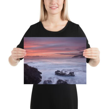 Una persona con una camisa negra sostiene una fotografía impresa del Póster rocas de Los Guirres, que captura bellamente una costa rocosa con olas rompiendo bajo un cielo de atardecer vibrante de color rosa y púrpura, que recuerda el encanto artístico que se encuentra en La Palma.