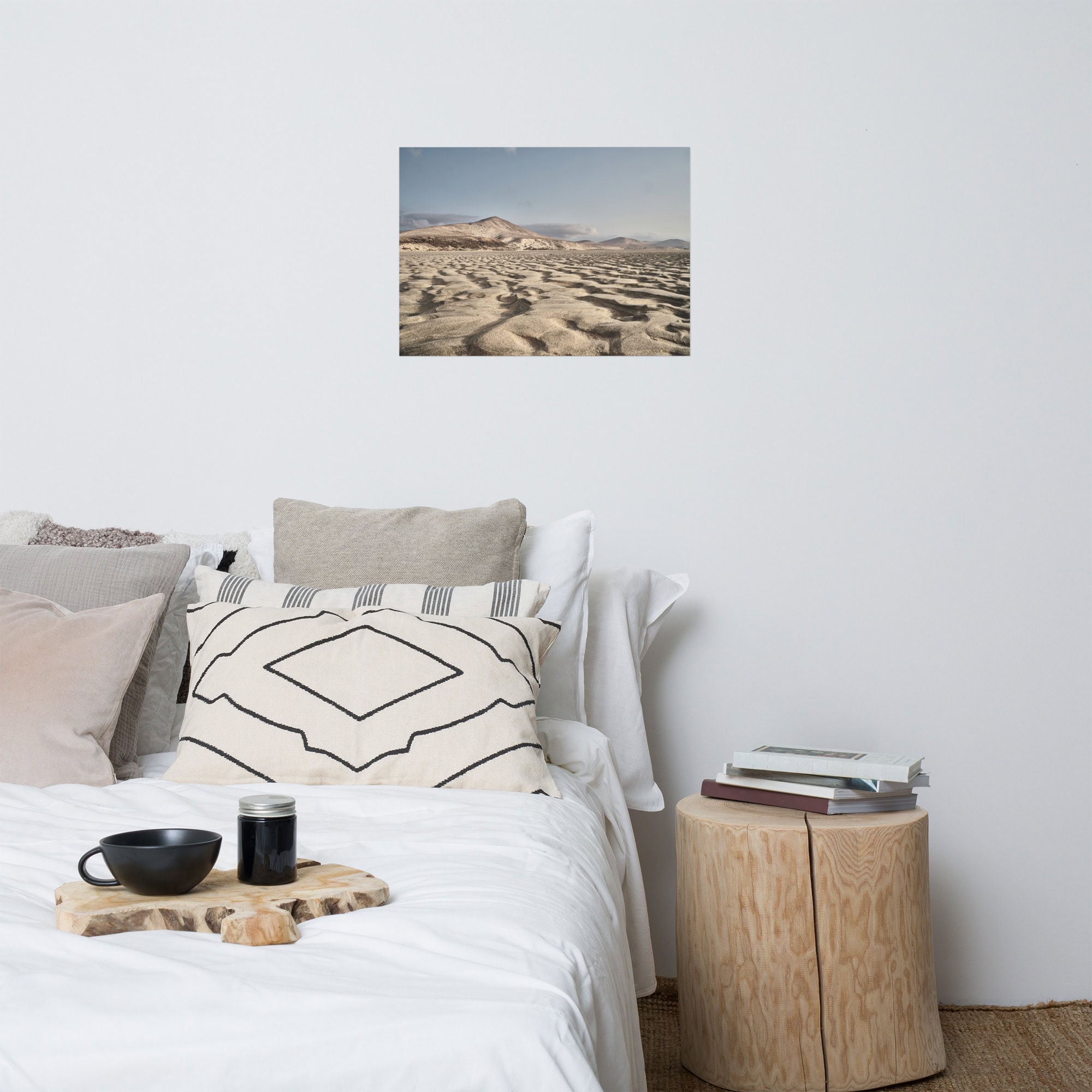 En un dormitorio minimalista, una cama cuidadosamente tendida está adornada con almohadas blancas y estampadas, acompañada de una mesilla de noche de madera con libros y una taza de La Palma. Sobre la cama cuelga el Póster Sotavento, que representa un paisaje desértico que complementa la gama cromática neutra con sus tonos tierra.