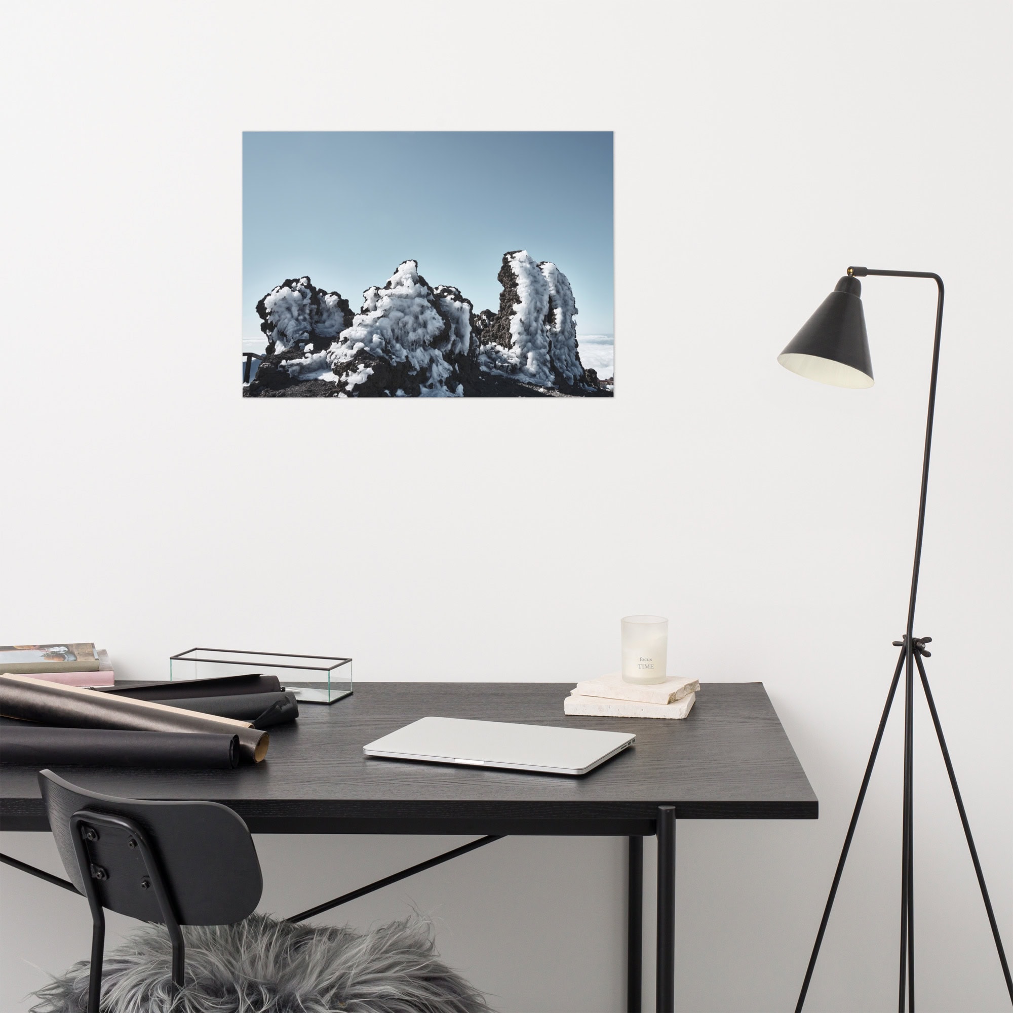 Un espacio de trabajo minimalista con un escritorio y una silla negros, realzados por Arte Decorativo. Un cojín peludo reposa sobre la silla. El escritorio cuenta con una computadora portátil, papeles enrollados y una vela. Una lámpara de pie negra se encuentra a un lado. La pared blanca está adornada con el póster Roque helado que muestra formaciones rocosas heladas.
