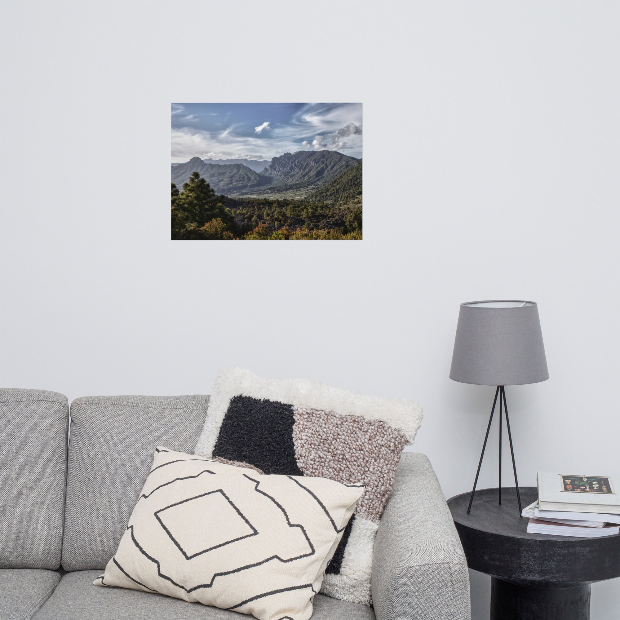 Una acogedora sala cuenta con un sofá gris con cojines estampados, perfecto para disfrutar de la belleza del arte decorativo. A su lado, una pequeña mesa redonda sostiene una lámpara gris. En la pared cuelga un exquisito "Póster Caldera de Taburiente", que muestra un impresionante paisaje de montañas y bosques, disponible en la tienda online como fotografías impresas.