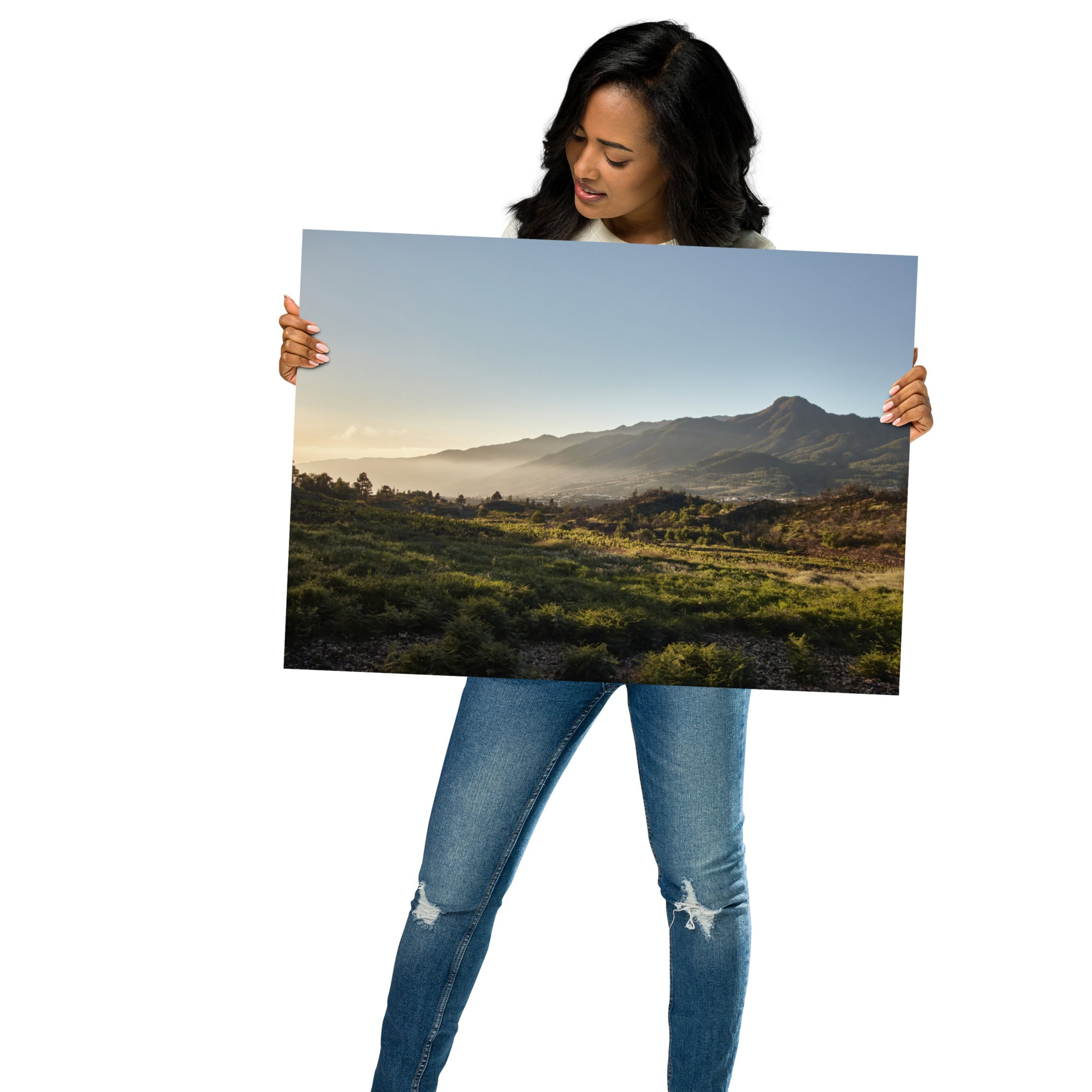 Una mujer con camisa blanca y jeans azules sostiene una gran fotografía impresa llamada "Póster atardecer desde Cuesta Juliana". Esta pieza de arte decorativo captura una pintoresca cadena montañosa bajo un cielo despejado, con la luz del sol proyectando sombras sobre campos de hierba, que recuerdan a los impresionantes paisajes canarios.