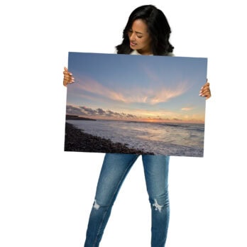 Vestido con jeans azules y una camisa oscura, un individuo sostiene una gran fotografía titulada "Póster atardecer en Los Guirres", que captura un tranquilo atardecer en la playa con nubes y olas, que encarna la esencia de Paisajes Canarios. El fondo blanco liso resalta esta pieza de Arte Decorativo.