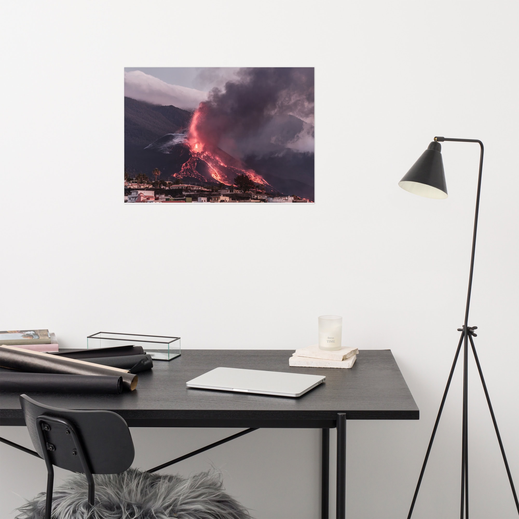 Un espacio de trabajo moderno muestra un escritorio negro con una computadora portátil cerrada, documentos, una vela y una silla forrada de piel. En la pared blanca sobre el escritorio cuelga un póster de Arte Decorativo titulado "Póster volcán 2021 La Palma edificios", que muestra una erupción volcánica espectacular con humo y lava. A un lado hay una lámpara de pie negra.
