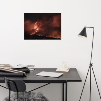 Un espacio de trabajo minimalista cuenta con un escritorio negro con una computadora portátil cerrada, cuadernos y una vela. Una silla negra con un cojín de piel gris está escondida debajo del escritorio. Cerca, hay una lámpara de pie negra en ángulo y un impresionante cartel "Póster volcán 2021 La Palma noche" adorna la pared.