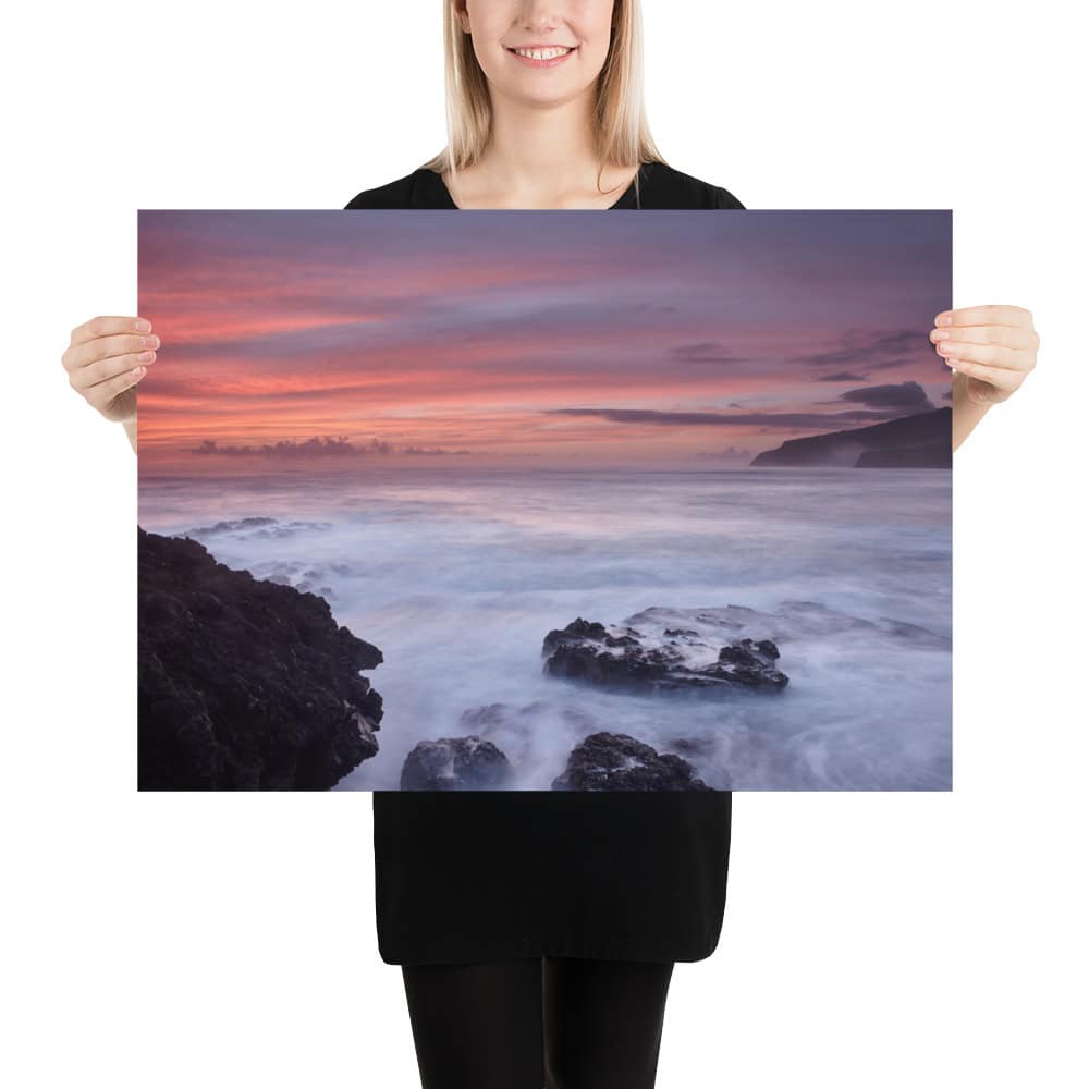 Una mujer sostiene un gran cartel titulado "Póster rocas de Los Guirres", que muestra una puesta de sol costera con olas rompiendo contra rocas oscuras bajo un cielo rosa y morado. Esta pieza es ideal para quienes buscan Arte Decorativo de una Tienda Online exclusiva, que recuerda a Paisajes Canarios.