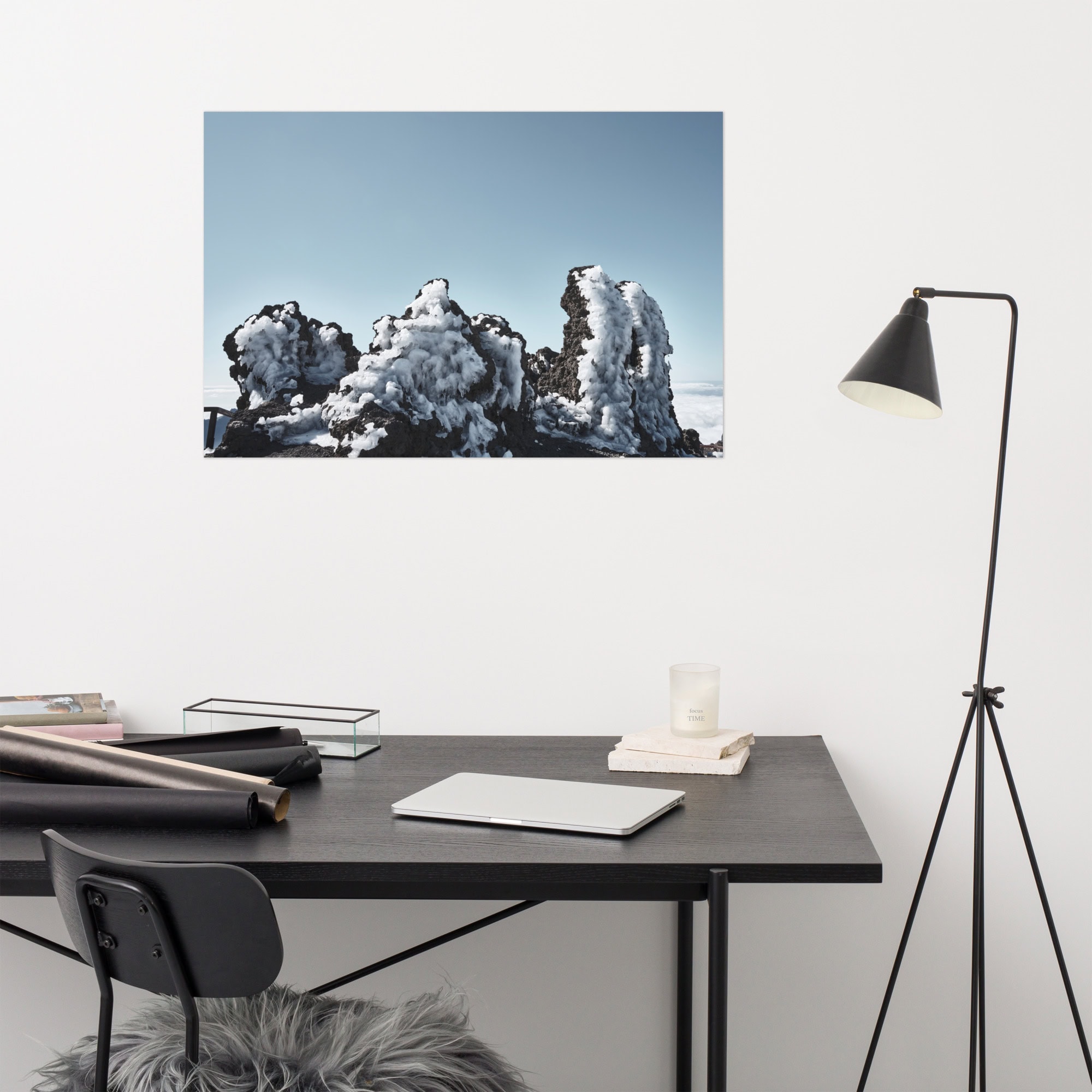 La oficina minimalista cuenta con un escritorio negro, una silla y una lámpara en ángulo. El escritorio está adornado con una computadora portátil, un cuaderno y una vela. El "Póster Roque helado" es una fotografía de rocas cubiertas de nieve bajo un cielo azul claro que realza su sensibilidad artística decorativa y le da un toque artístico al espacio.