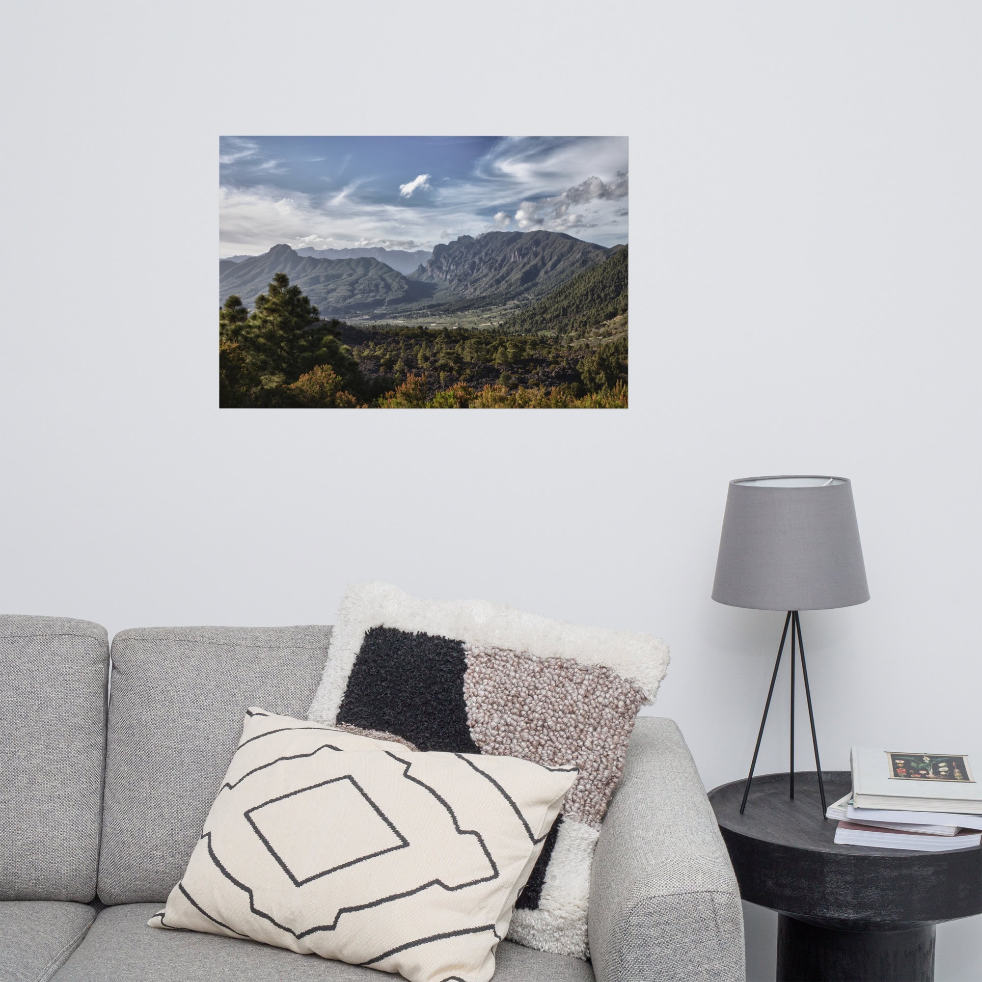 En la sala de estar hay un sofá gris adornado con cojines estampados, complementado por una mesa auxiliar redonda que sostiene una lámpara gris. Sobre el sofá cuelga el póster Caldera de Taburiente de Paisajes Canarios, que representa un paisaje montañoso que añade encanto a la pared blanca.