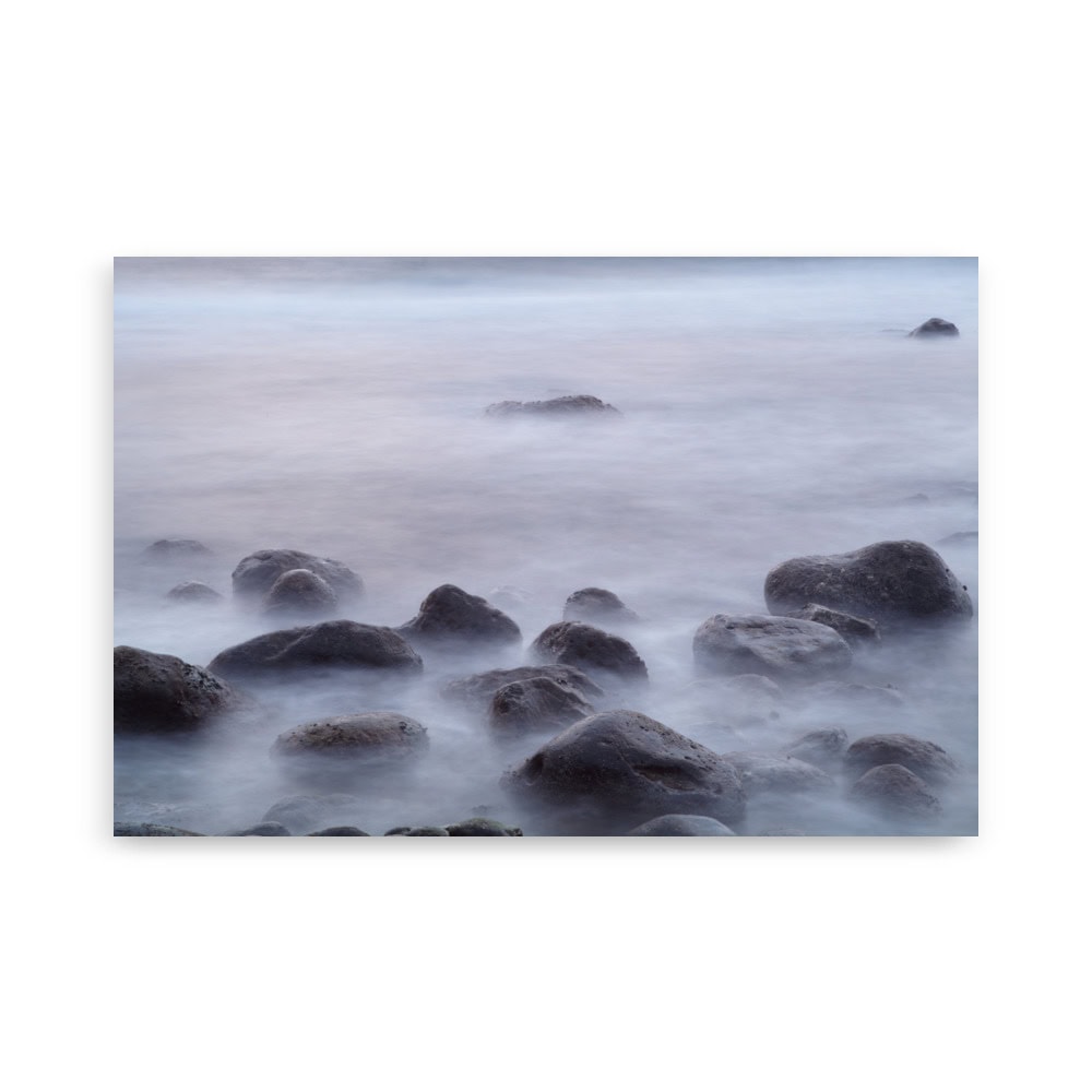 El cartel de rocas de la playa de El Lloret muestra un paisaje marino sereno cerca de La Palma, con grandes rocas cubiertas de musgo parcialmente sumergidas en aguas tranquilas y brumosas. El cielo suavemente iluminado realza la atmósfera tranquila y etérea, que evoca el arte decorativo.