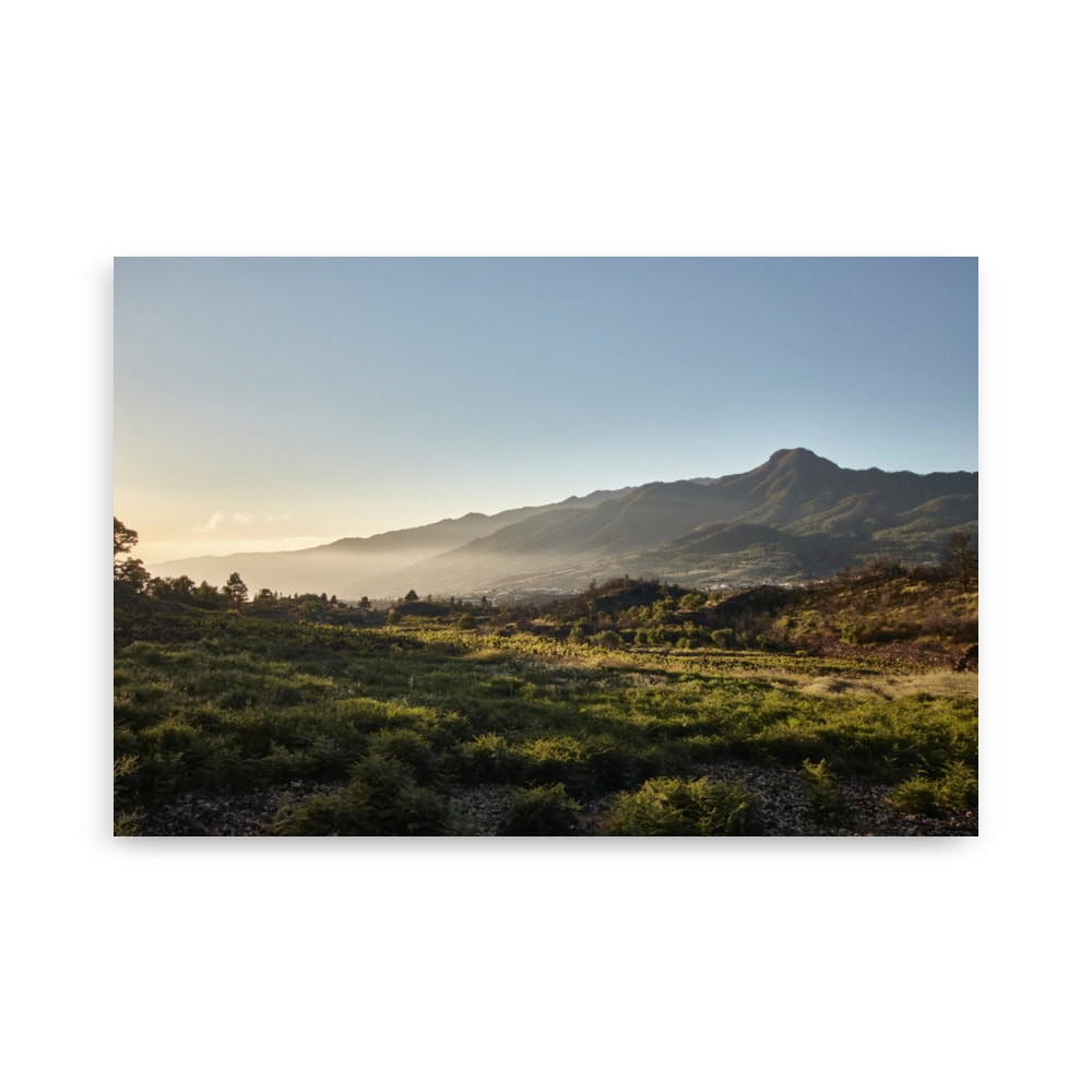 Un paisaje espectacular con un campo de hierba en primer plano y una cadena montañosa bajo el cielo azul claro, que captura la esencia de la belleza de La Palma. La luz del sol ilumina esta escena serena, lo que la hace ideal para capturarla en nuestro "Póster atardecer desde Cuesta Juliana", disponible a través de nuestra tienda online.