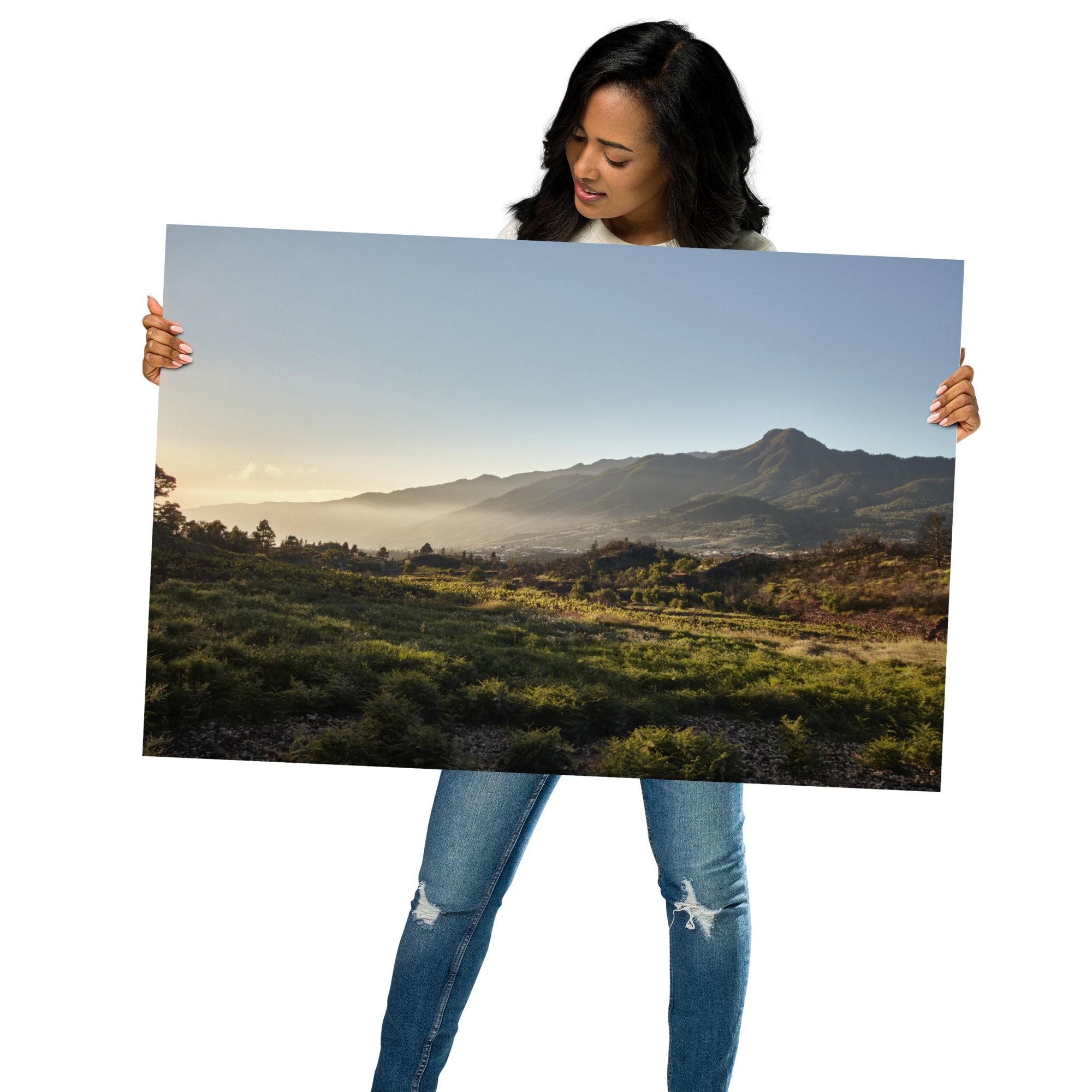 Una persona sosteniendo el Póster atardecer desde Cuesta Juliana, ideal para Arte Decorativo, muestra un campo verde exuberante y montañas distantes bajo un cielo azul claro. Vestido con jeans azules, el individuo admira esta vibrante Fotografía Impresa.
