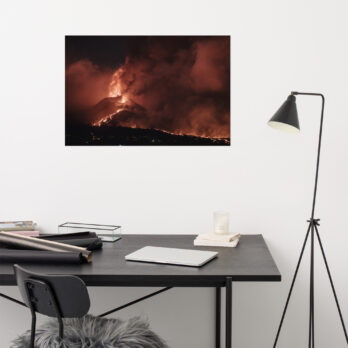 Un espacio de trabajo minimalista que muestra un escritorio negro adornado con una computadora portátil, una pila de papeles y una vela. Sobre el escritorio cuelga el vívido "Póster volcán 2021 La Palma noche", que muestra un volcán en erupción con lava brillante, disponible en nuestra tienda en línea. Cerca hay una lámpara de pie moderna.