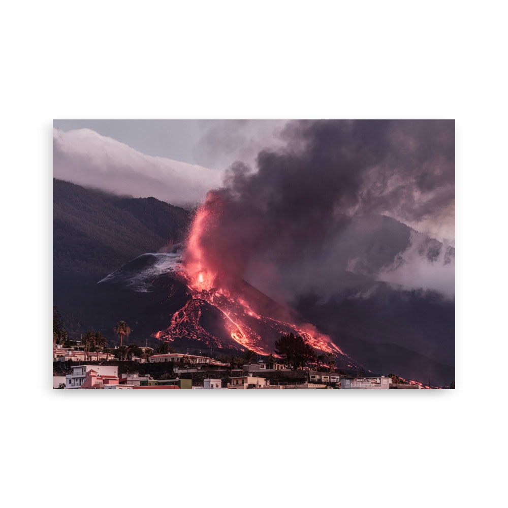 Una erupción volcánica proyecta lava y cenizas hacia el cielo en medio de una nube de humo oscuro. La lava cae en cascada por la montaña, creando una escena impactante que refleja la esencia del Póster volcán 2021 La Palma edificios, con casas que aparecen en primer plano bajo un cielo nublado.