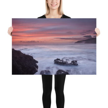 Una persona sostiene un gran cartel titulado "Póster rocas de Los Guirres", que muestra un sereno paisaje marino de La Palma con una vibrante puesta de sol. El cielo está pintado en tonos rosa y naranja, mientras las olas chocan suavemente contra las oscuras rocas costeras, que recuerdan a las fotografías impresas de alta calidad disponibles en nuestra tienda online.