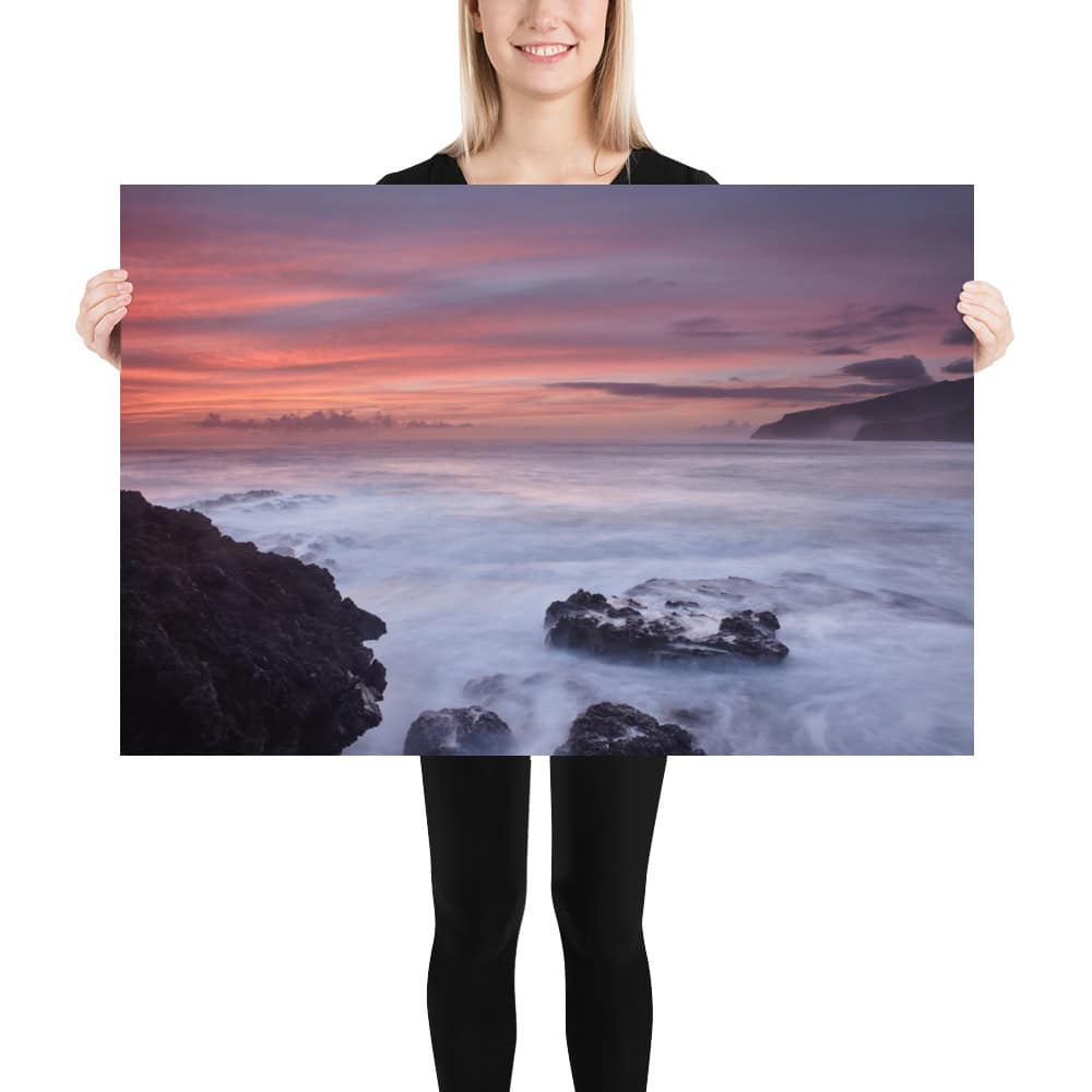 Una persona sostiene un gran cartel titulado "Póster rocas de Los Guirres", que muestra un sereno paisaje marino de La Palma con una vibrante puesta de sol. El cielo está pintado en tonos rosa y naranja, mientras las olas chocan suavemente contra las oscuras rocas costeras, que recuerdan a las fotografías impresas de alta calidad disponibles en nuestra tienda online.