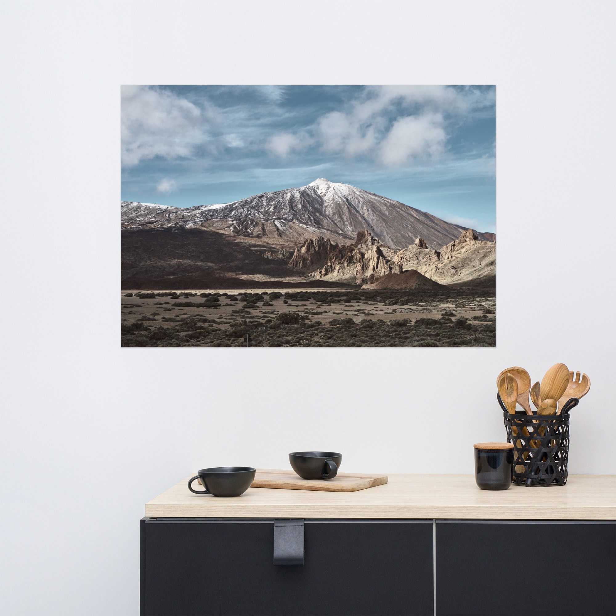 El póster Teide, que muestra un sereno paisaje montañoso con un pico nevado bajo un cielo parcialmente nublado, se exhibe sobre una encimera de cocina minimalista con un cajón negro. Sobre la encimera hay dos cuencos negros y un soporte también negro lleno de utensilios de madera.