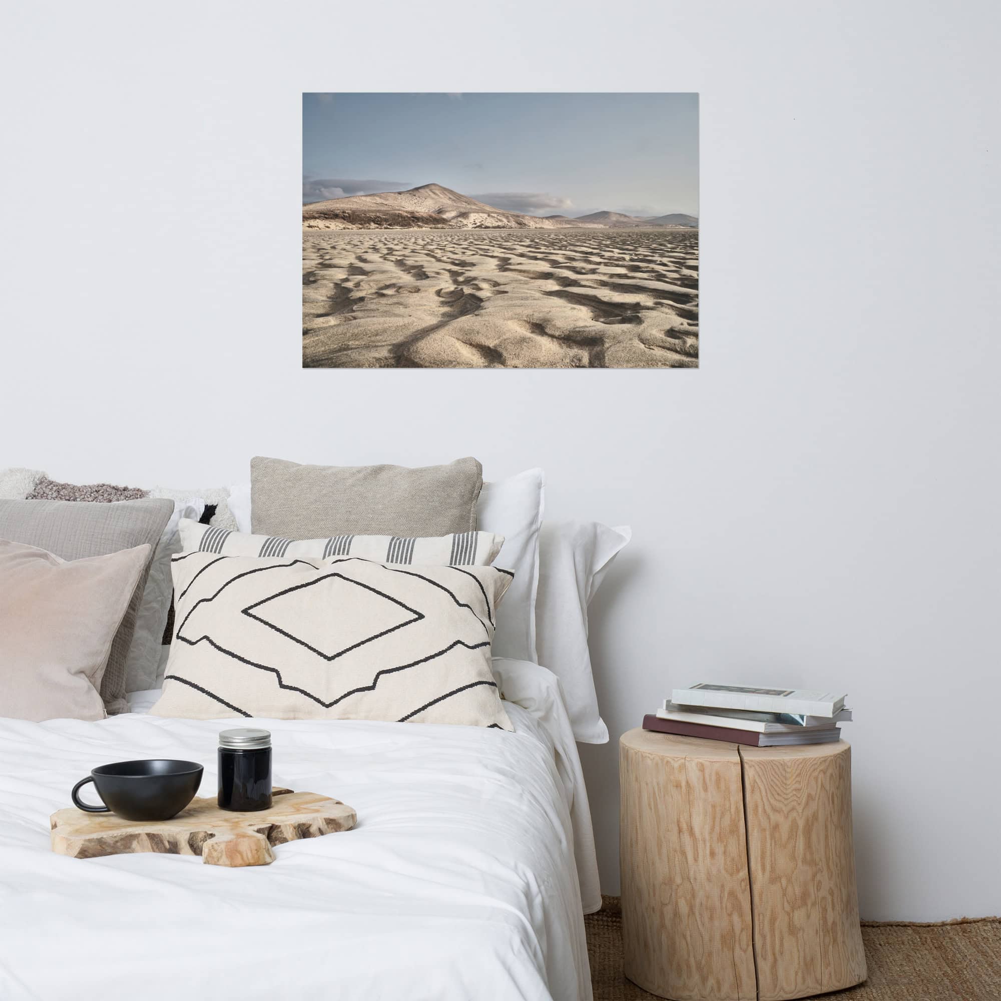 Un dormitorio acogedor muestra una cama blanca cuidadosamente hecha y adornada con almohadas estampadas, una mesa auxiliar de madera con libros y una taza, y una encantadora fotografía de un paisaje desértico en la pared sobre la cama. Agregue calidez explorando Tienda Online para encontrar el hermoso "Póster Sotavento" para realzar su espacio.