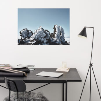 Un espacio de trabajo minimalista cuenta con un escritorio negro con un ordenador portátil, papeles enrollados y una vela. Cerca hay una lámpara de pie moderna. En la pared blanca cuelga una impresionante pieza de arte decorativo: el póster Roque helado, que muestra rocas cubiertas de nieve bajo un cielo azul claro.