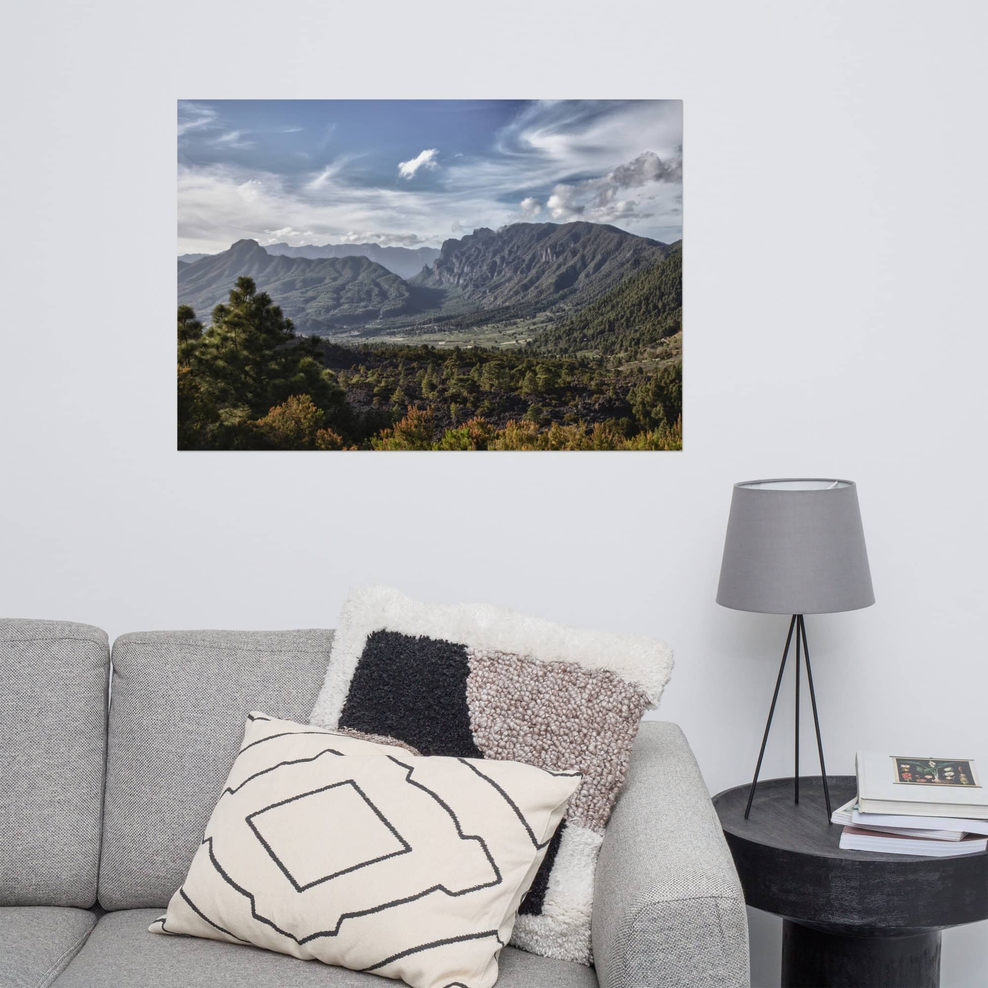 Un póster de Caldera de Taburiente de Volcanica Productions cuelga sobre una pared blanca encima de un sofá gris. La imagen muestra unas montañas verdes y exuberantes bajo un cielo parcialmente nublado. Junto al sofá hay una mesa auxiliar negra adornada con una lámpara gris y una almohada estampada, disponible en nuestra Tienda Online.