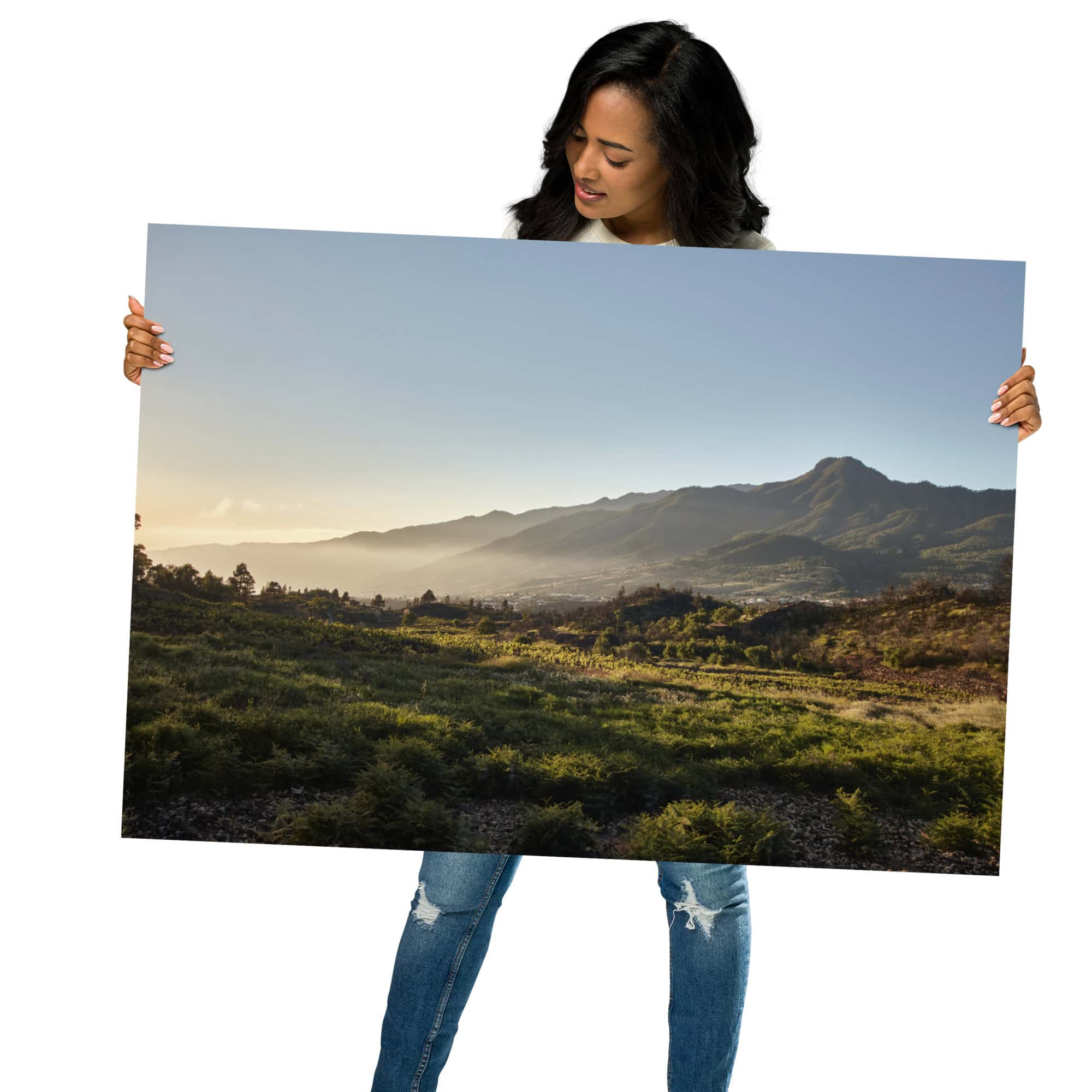 Una mujer con una camiseta negra y unos vaqueros azules muestra el "Póster atardecer desde Cuesta Juliana", disponible en nuestra tienda online. Esta exquisita pieza de arte decorativo captura una vista panorámica de un campo de hierba con montañas distantes bajo un cielo azul, perfecta para quienes aprecian las fotografías impresas de calidad.