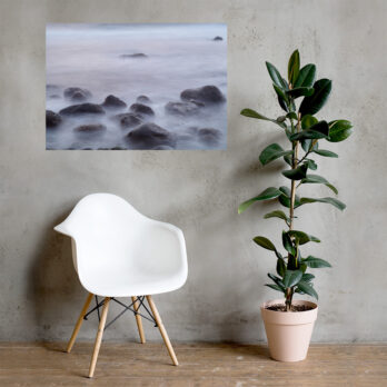 Un interior moderno con una silla blanca y una planta en maceta sobre un suelo de madera contra una pared gris. Sobre la silla, una exquisita pieza de arte mural titulada "Póster de Rocas de la playa de El Lloret" de Paisajes Canarios captura una escena serena de rocas envueltas en agua brumosa, agregando un toque artístico que recuerda a los tranquilos paisajes de La Palma.