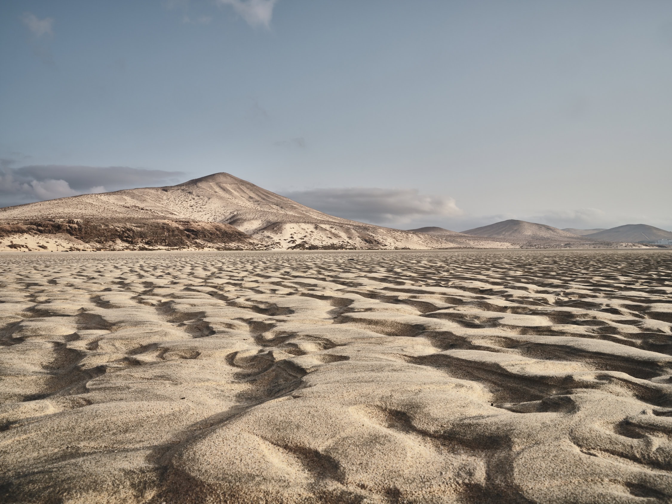 Un vasto paisaje desértico con onduladas dunas de arena bajo un cielo azul claro. Al fondo, varios picos de montañas se elevan majestuosos, creando una escena digna de un Póster Sotavento, que encarna el Arte Decorativo sereno y expansivo.