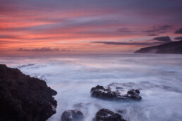 Una tranquila vista costera de Los Guirres al atardecer muestra una costa rocosa con olas que acarician suavemente las rocas. El cielo está adornado con tonos de rosa, naranja y violeta, con acantilados recortados en el horizonte, una captura ideal para Póster rocas de Los Guirres como una exquisita pieza de arte decorativo.