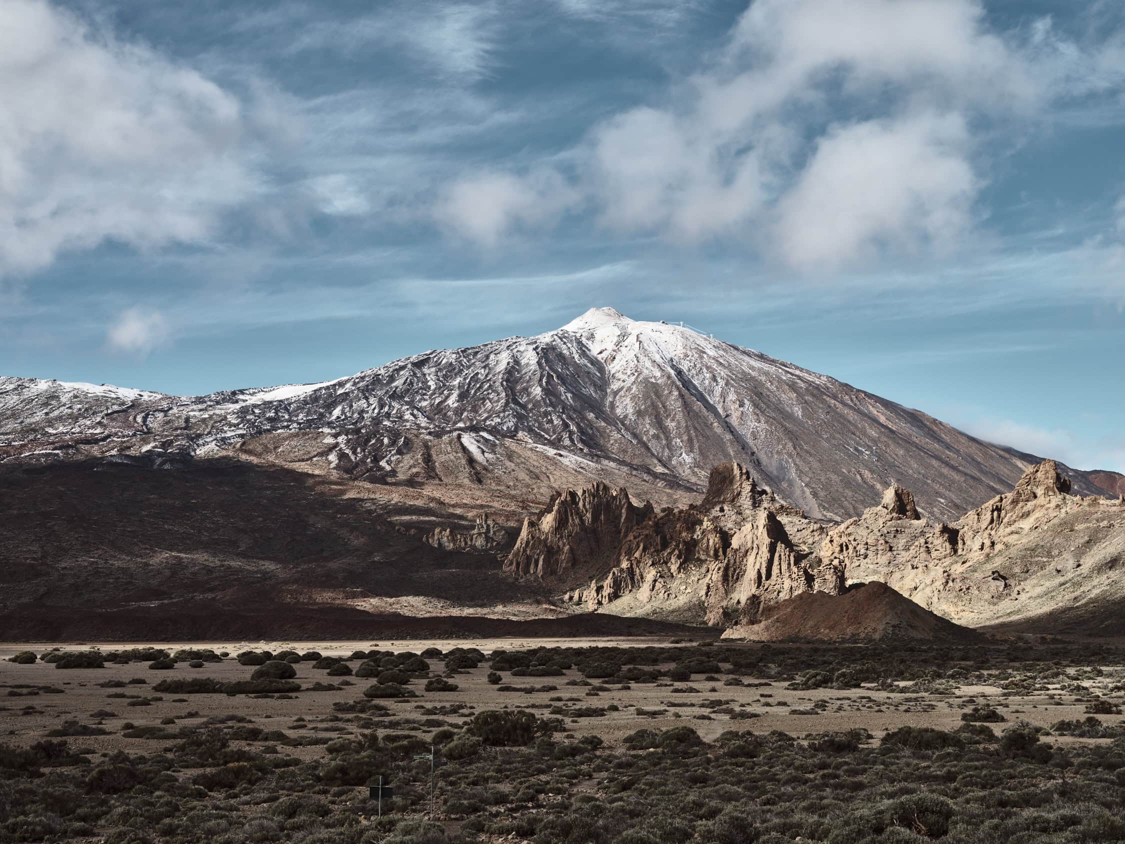 Una vista panorámica de una montaña cubierta de nieve bajo un cielo parcialmente nublado muestra el arte de la naturaleza que recuerda a una obra maestra del arte decorativo. En primer plano se ven afloramientos rocosos y un paisaje desértico, donde las escarpadas laderas de la montaña contrastan maravillosamente con el terreno extenso y llano.