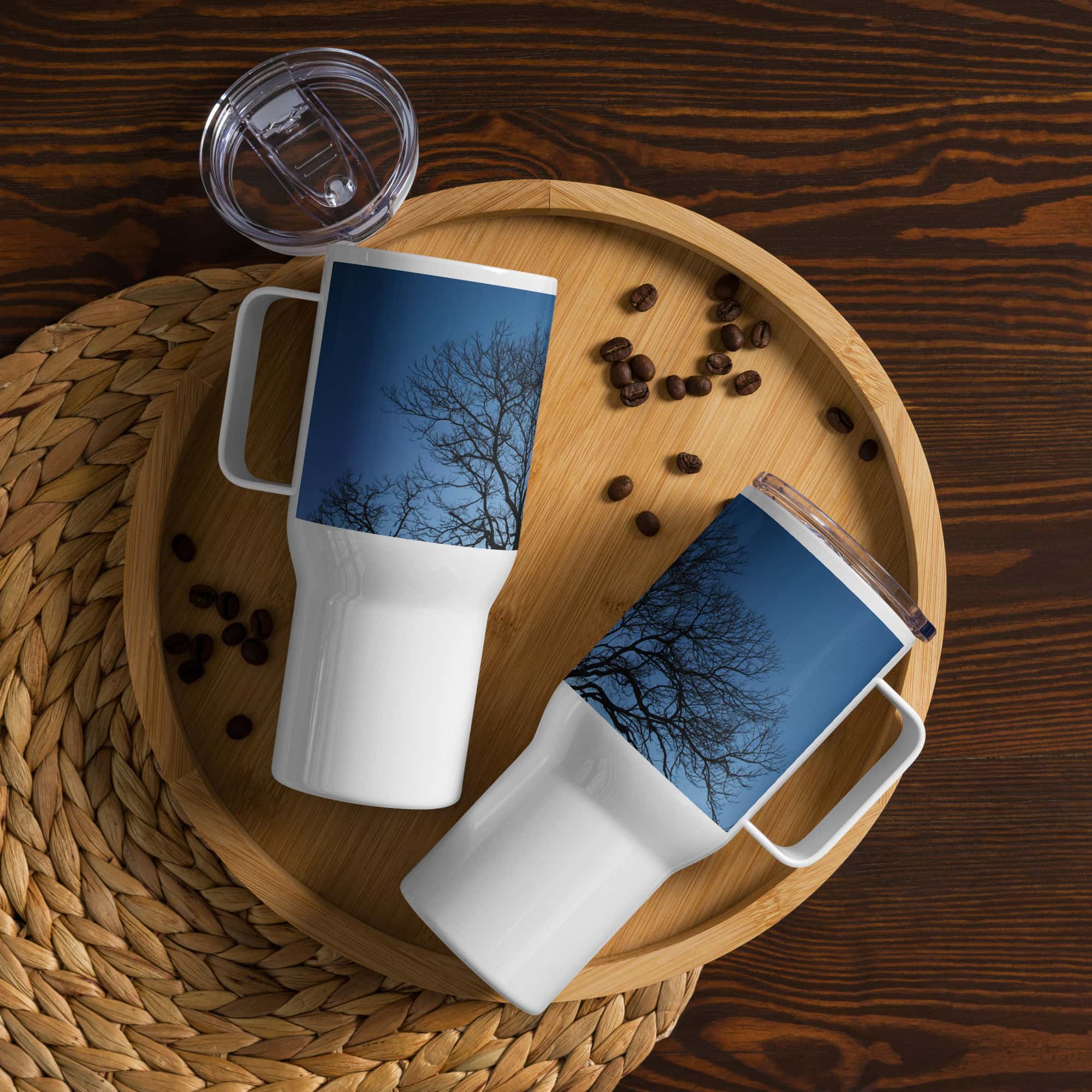 Dos tazas térmicas blancas "Termo con asa ramas fondo azul" con un diseño de ramas de árbol se exhiben en una bandeja de madera, rodeadas de granos de café dispersos. Cerca hay una tapa colocada sobre una estera tejida sobre una superficie de madera oscura. Estas piezas artísticas reflejan el encanto de Paisajes Canarios, ahora disponibles en nuestra tienda online.