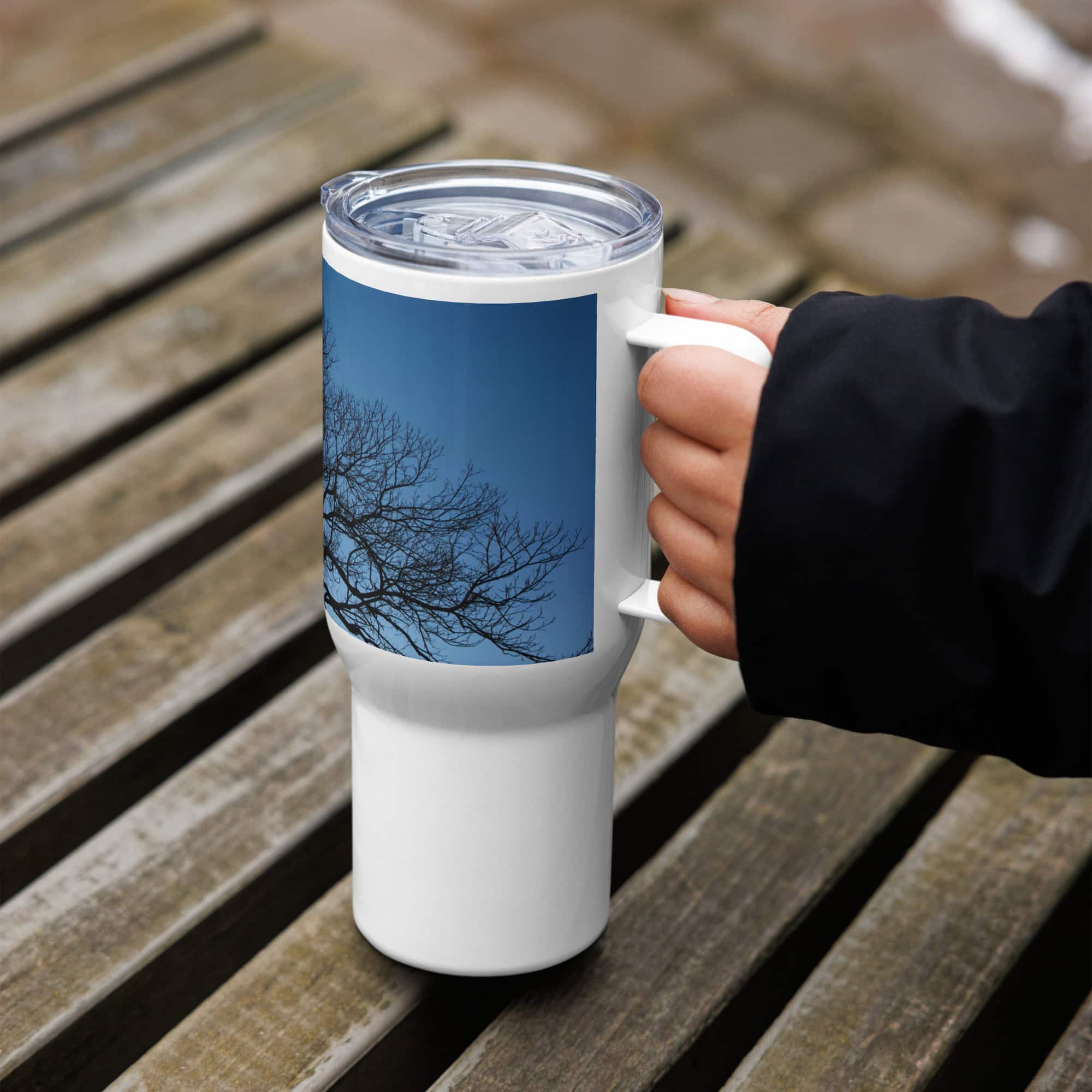 Una persona vestida con una manga negra sostiene el "Termo con asa ramas fondo azul", una taza de viaje blanca de Volcanica Productions que muestra un árbol desnudo contra un cielo azul claro. Disponible en su tienda en línea, la taza está colocada sobre un banco de madera con diseño de listones, que captura el encanto natural de La Palma.