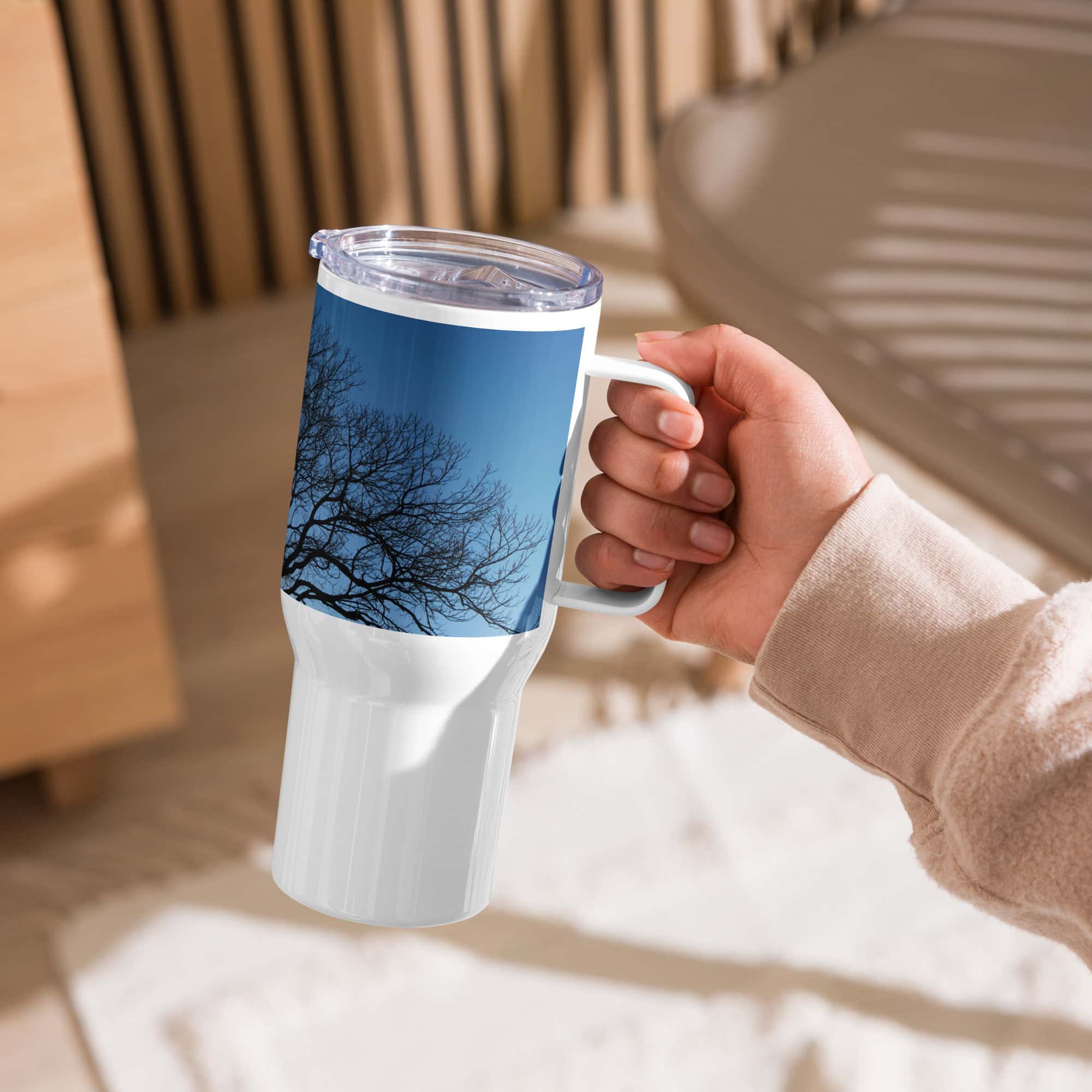 Una mano sostiene el "Termo con asa ramas fondo azul", una taza de viaje blanca que muestra la foto de un árbol sin hojas contra un cielo azul, lo que la convierte en un regalo perfecto. El ambiente interior con muebles de madera y la luz del sol que proyecta sombras crea una atmósfera acogedora, que refleja la atención al detalle de Volcanica Productions.