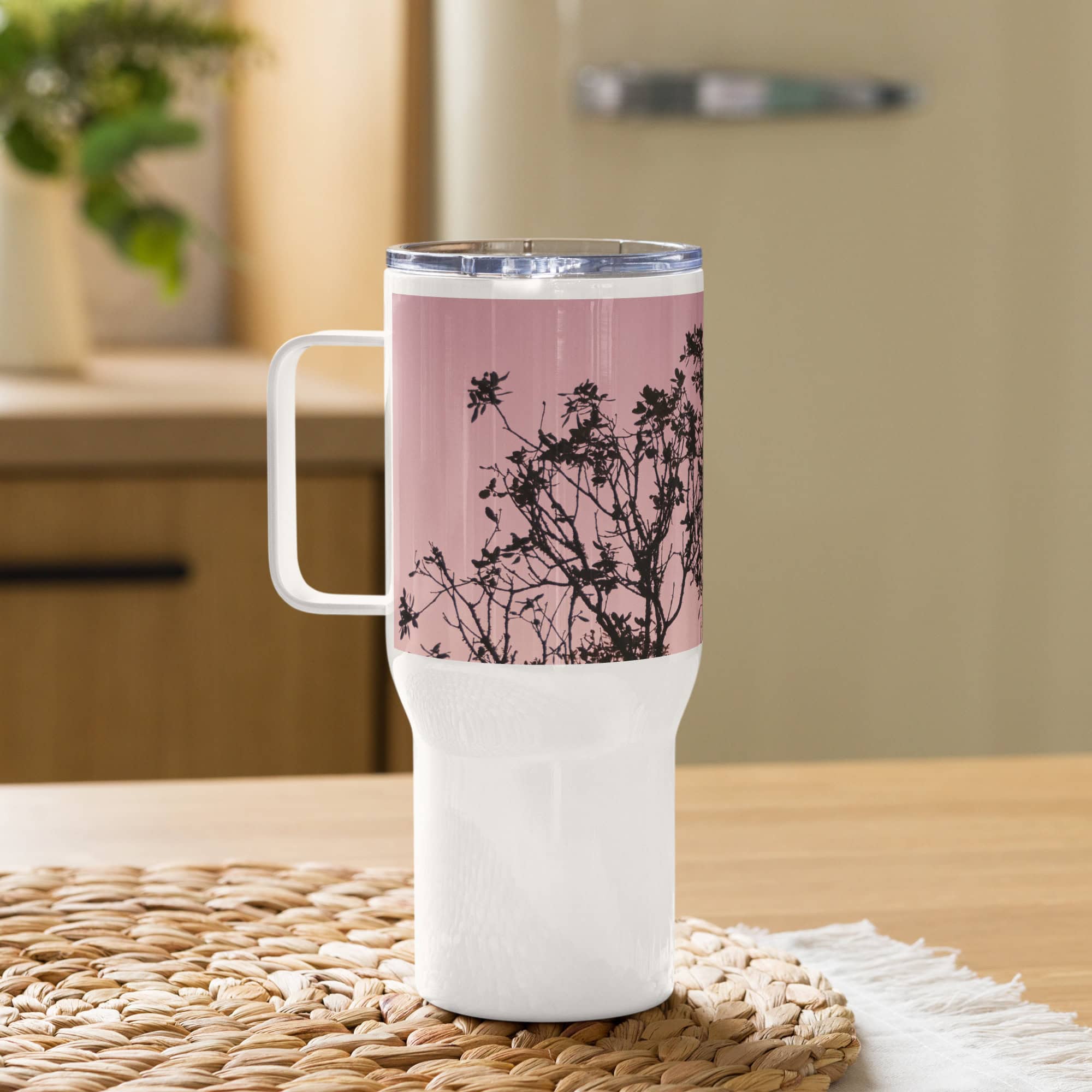El "Termo con asa ramas fondo rosa" es una taza de viaje alta de color blanco que presenta una tapa rosa y un elegante diseño de ramas florales. Se apoya sobre una estera tejida colocada sobre una superficie de madera, lo que la convierte en el "regalo" perfecto para cualquier persona. La taza combina a la perfección con fondos de cocina suavemente difuminados y vegetación sutil.