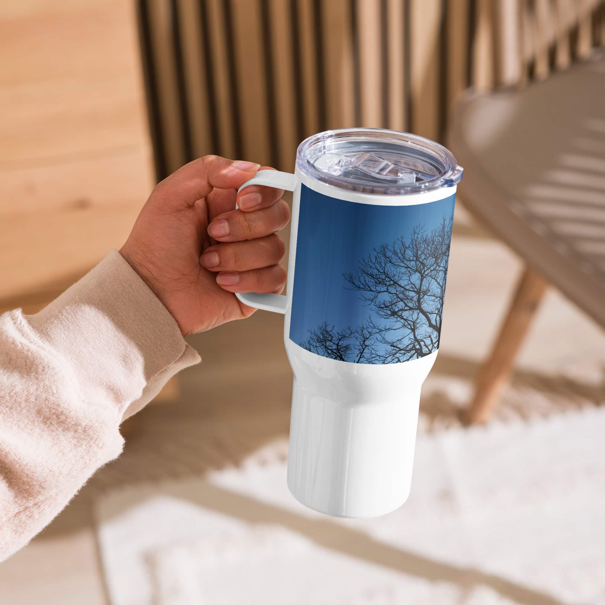 Una persona sostiene un Termo con asa ramas fondo azul, con una imagen de los árboles de La Palma con ramas desnudas contra un cielo azul vibrante. En el fondo, una silla de madera borrosa y una alfombra beige crean un ambiente acogedor, ideal para comprar regalos en línea o para los fanáticos de los regalos únicos.