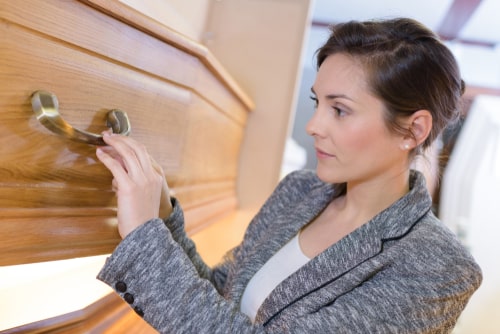 A female funeral director in a gray suit adjusts the handle of a light-toned oak coffin.