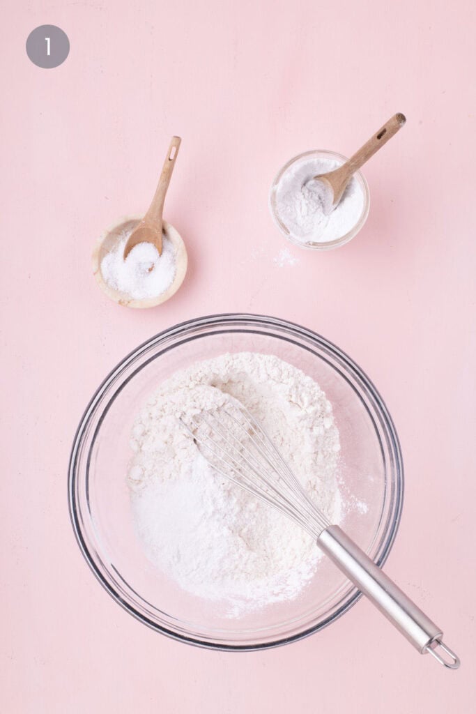Flour, baking soda, and salt whisked together in a glass bowl on a pink background for cornflake cookie dough.