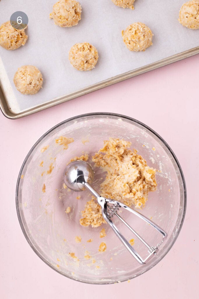 Scooped cornflake cookie dough on a parchment-lined baking sheet, with a bowl of remaining dough and a cookie scoop nearby.