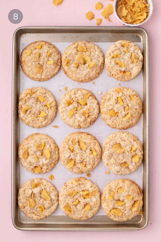 Freshly baked cornflake cookies on a parchment-lined baking sheet with extra cornflakes scattered nearby.
