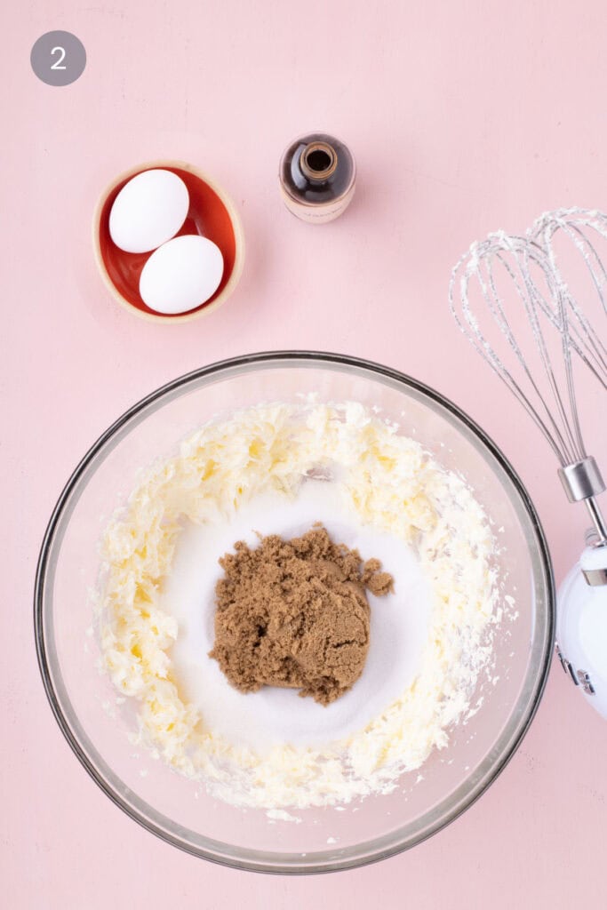 A glass bowl filled with creamed butter topped with granulated sugar, brown sugar, next to two eggs, a bottle of vanilla extract, and a hand mixer on a pink background.