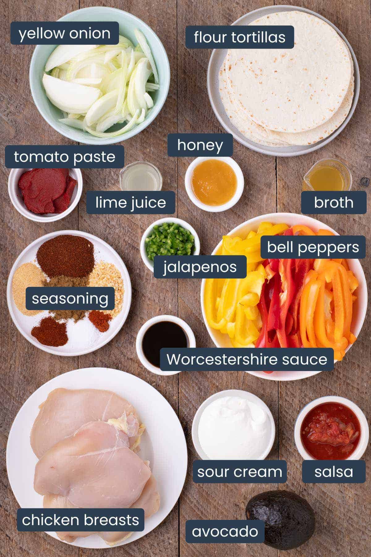 Flat lay of slow cooker chicken fajitas ingredients including raw chicken breasts, sliced bell peppers, onions, garlic, chilli powder, garlic powder, onion powder, cumin, smoked paprika, cayenne pepper, Worcestershire sauce, lime juice, honey, salsa, tortillas, sour cream, avocado, and other sauces arranged on a rustic wooden table.