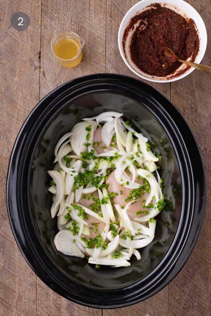 Raw chicken breasts layered with sliced onions and jalapeños in a slow cooker, with broth and spice paste off to the side.