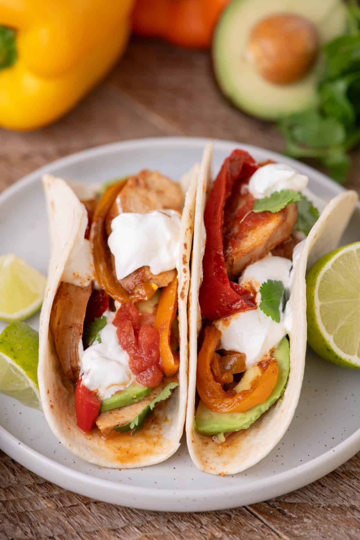 Two soft tortillas filled with sliced chicken, sautéed bell peppers, avocado, sour cream, and salsa, served with lime wedges on a rustic plate.