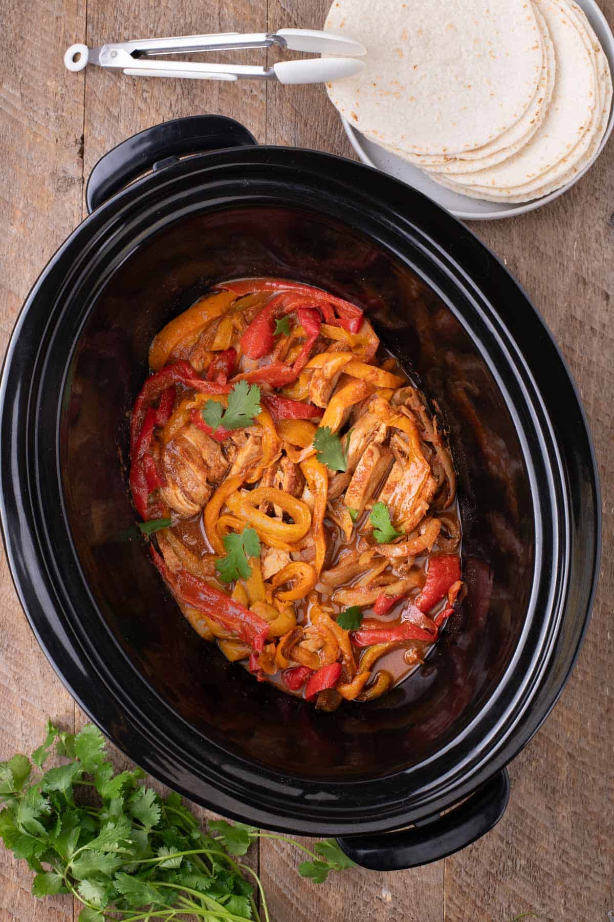 Overhead view of shredded chicken and sautéed bell peppers in a slow cooker, garnished with fresh cilantro, with tortillas and tongs on the side.