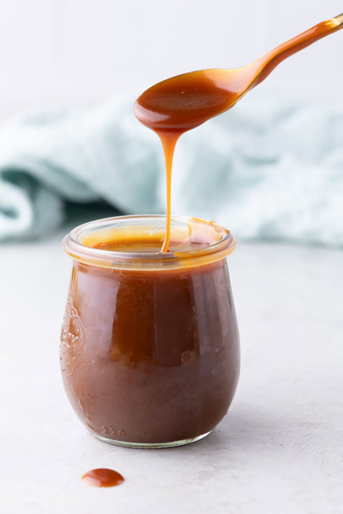 Golden spoon lifting salted caramel sauce over a glass jar with caramel dripping back i