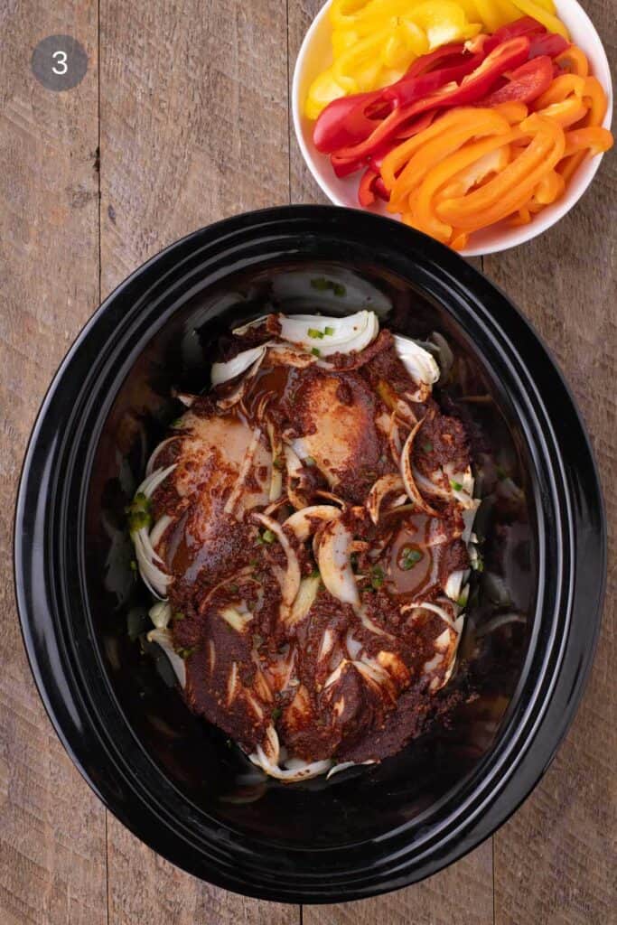 Slow cooker filled with seasoned chicken and onions coated in a deep red spice paste, peppers set aside nearby.