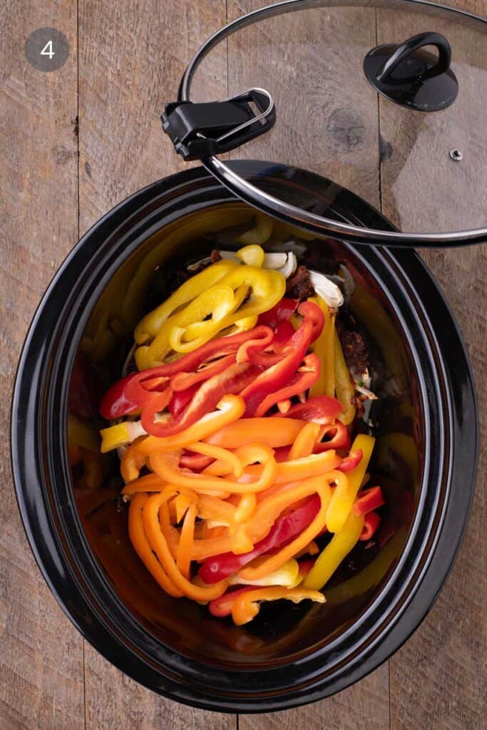 Sliced red, yellow, and orange bell peppers covering the chicken in a slow cooker, lid placed and ready to cook.
