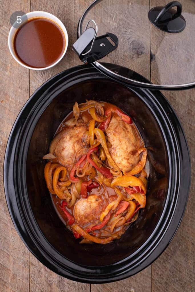 Juicy cooked chicken and vegetables resting in their own rich liquid inside the slow cooker, with a small bowl of sauce set aside.
