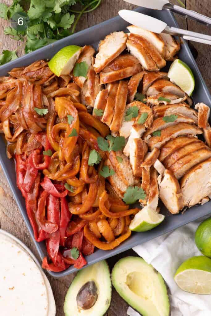 Sliced slow-cooked chicken and sautéed red, orange, and yellow peppers arranged on a serving tray with lime wedges, cilantro, avocado halves, and soft tortillas on the side.