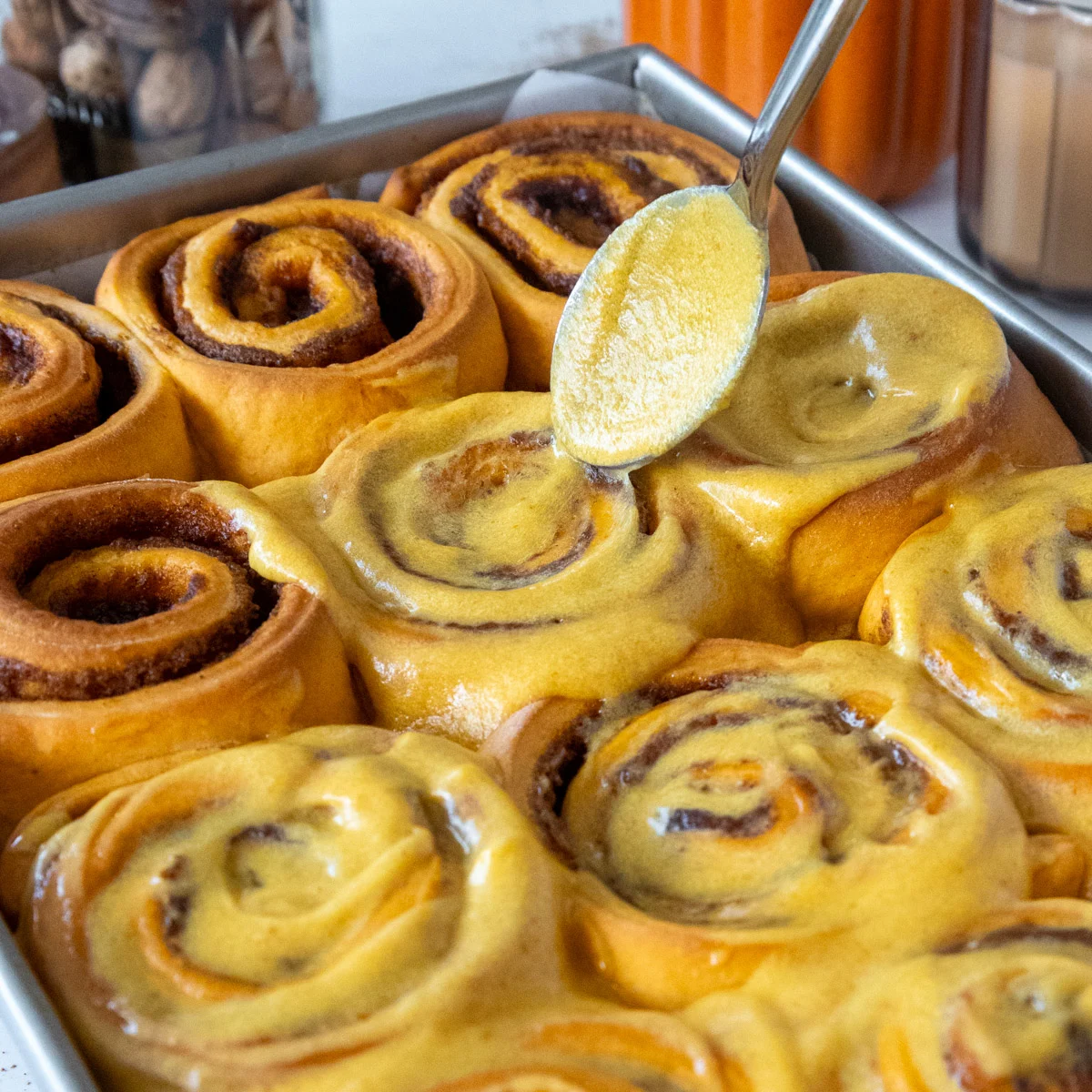 image of pumpkin pie cinnamon rolls being frosted with pumpkin cream cheese frosting